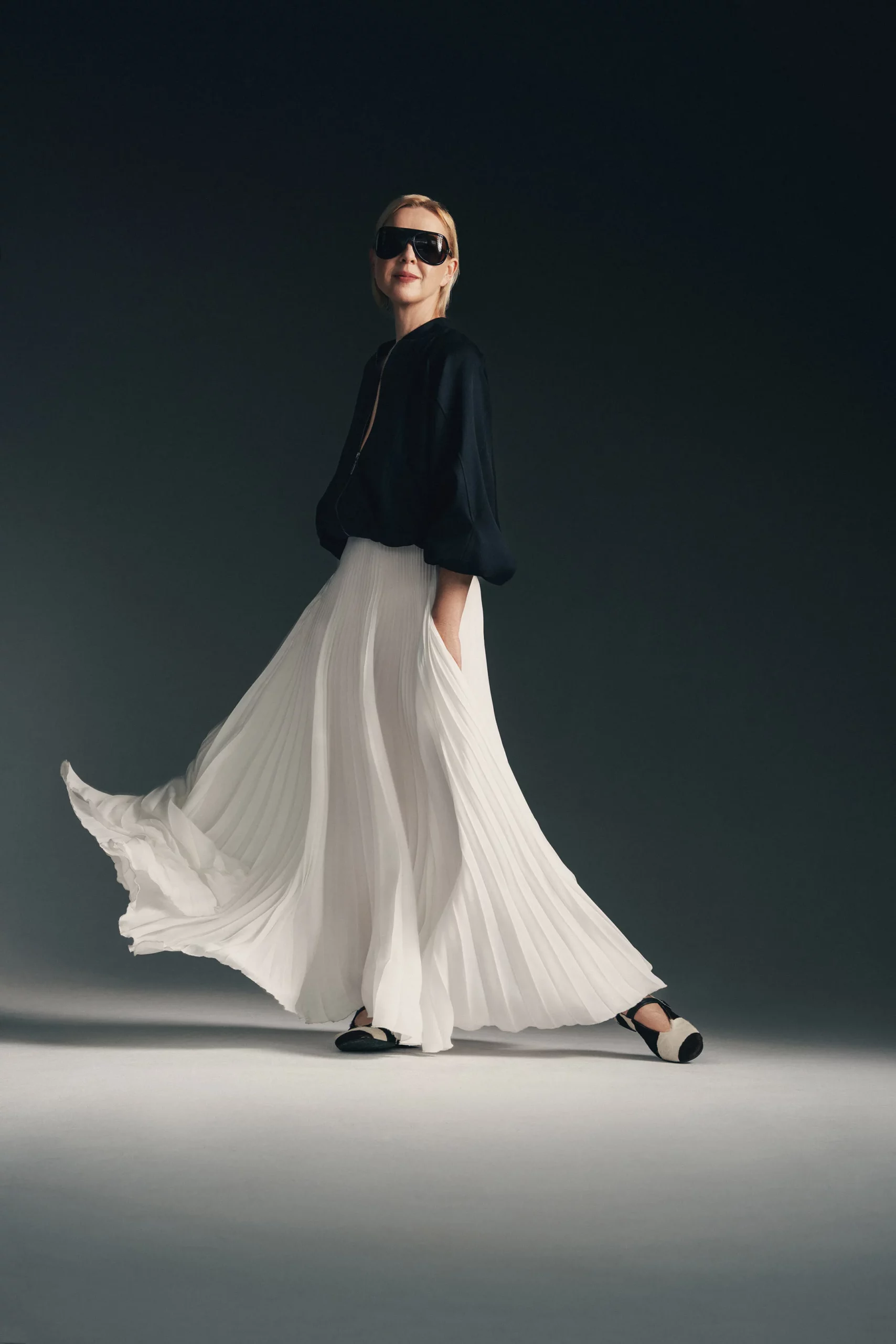 Modelka w długiej plisowanej spódnicy Soleil marki JAK, uszytej z lekkiej, falującej tkaniny, siedzi w minimalistycznej scenerii