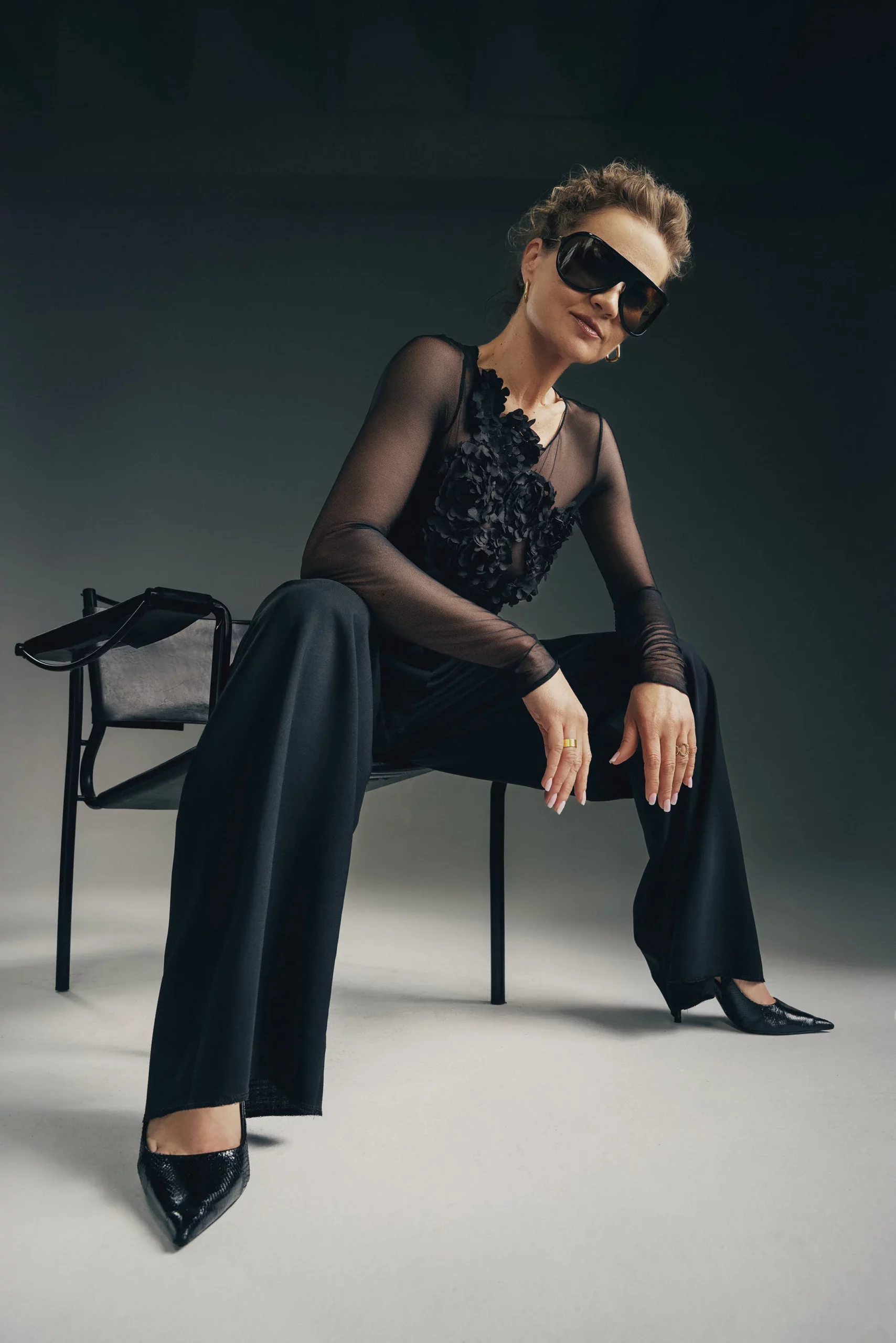 Kobieta w eleganckim komplecie – przezroczyste body z kwiatowymi aplikacjami 3D i czarne, szerokie spodnie, siedzi na designerskim krześle w studiu