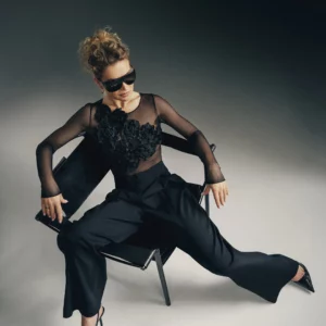 Kobieta w eleganckim komplecie – przezroczyste body z kwiatowymi aplikacjami 3D i czarne, szerokie spodnie, siedzi na designerskim krześle w studiu
