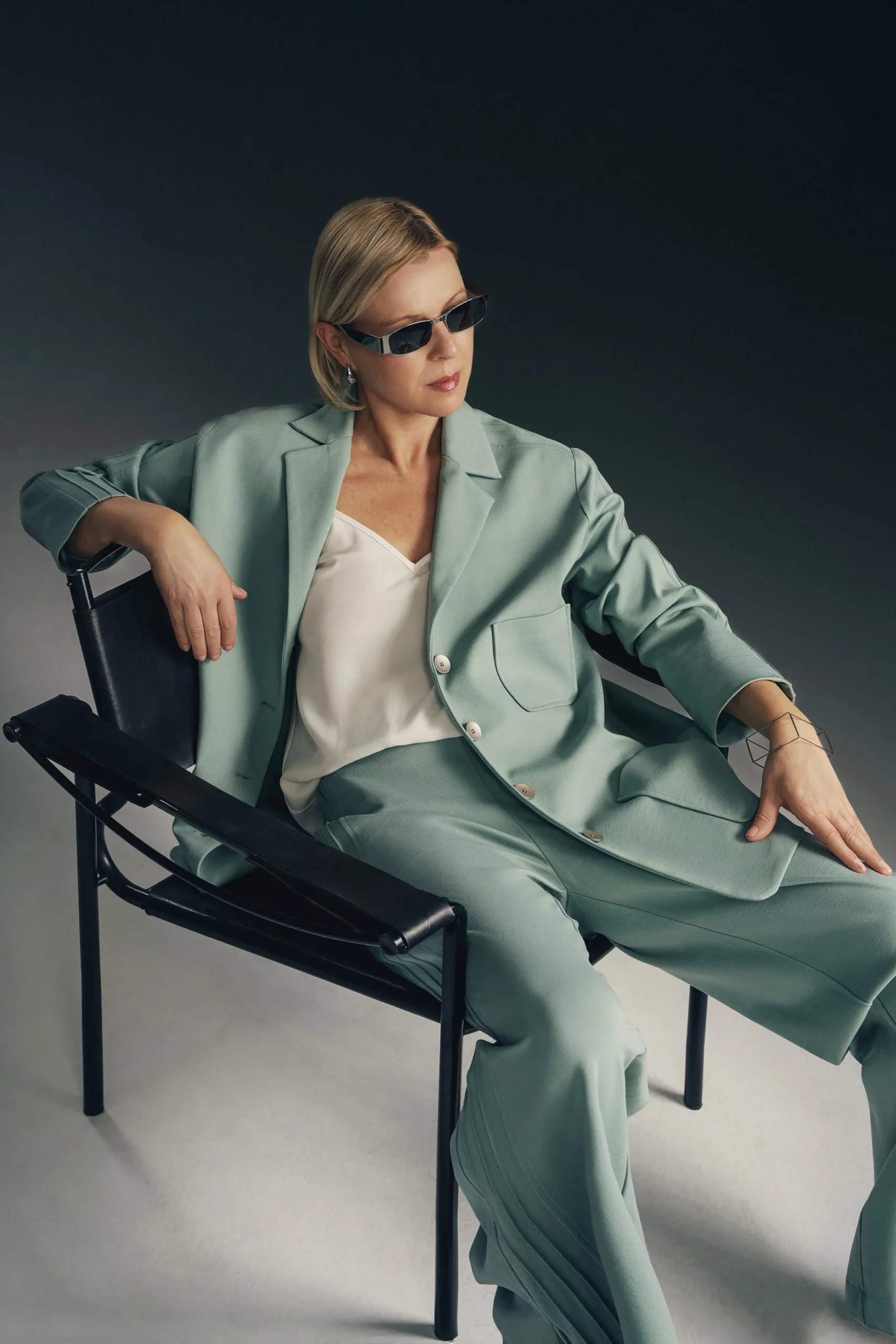 Kobieta siedzi na czarnym krześle, ubrana w zestaw w kolorze szałwi - luźną oversizową marynarkę Jazz z ozdobnymi lampasami na rękawach i dopasowane do marynarki spodnie. Pod marynarką ma biały top na ramiączkach. Stylizacja jest nowoczesna, elegancka i utrzymana w sportowej elegancji