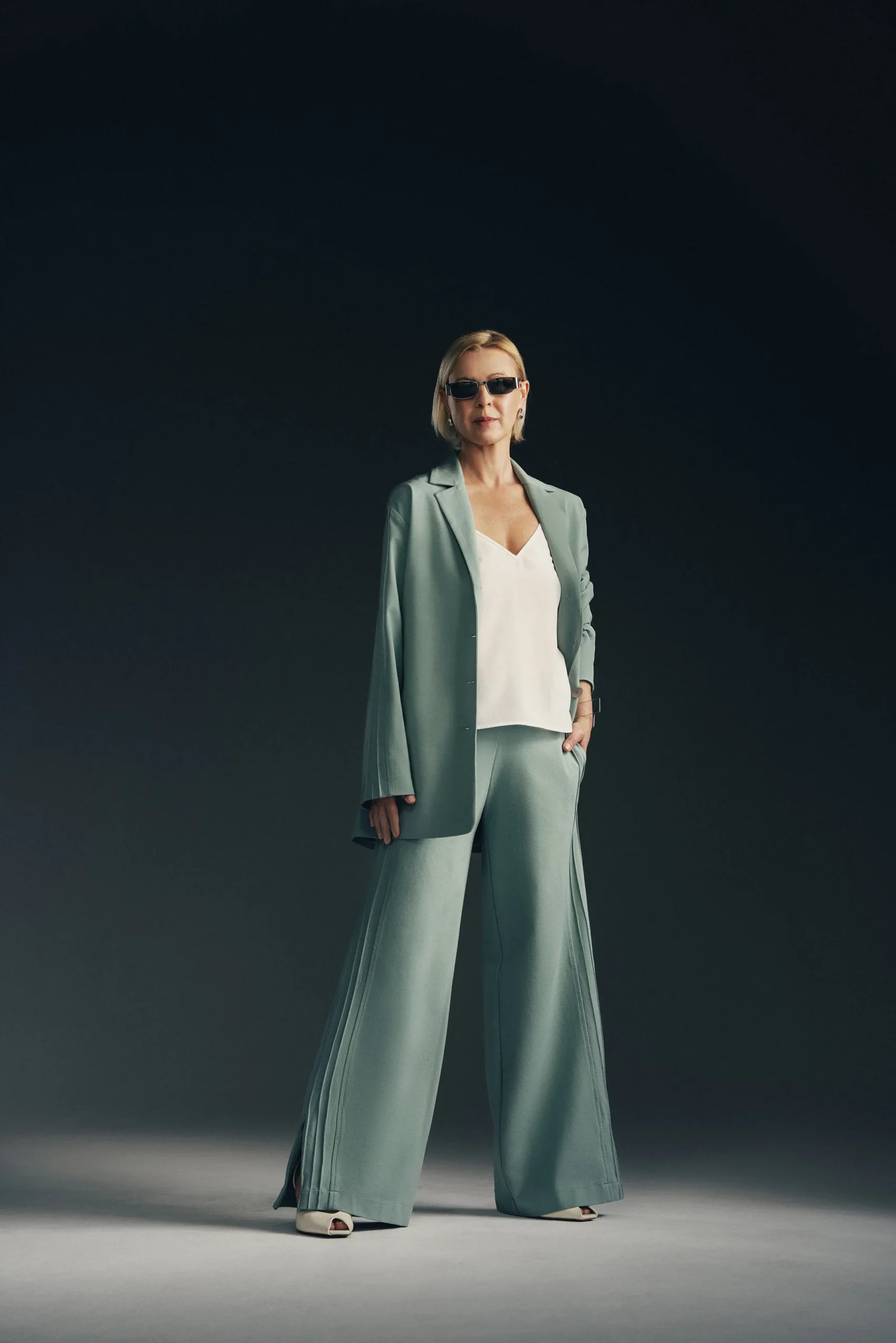 Kobieta ubrana w zestaw w kolorze szałwi z nutą mięty- luźną oversizową marynarkę Jazz z ozdobnymi lampasami na rękawach i dopasowane do marynarki spodnie. Pod marynarką ma biały top na ramiączkach. Stylizacja jest nowoczesna, elegancka i utrzymana w sportowej elegancji. Spodnie jazz z ozdobnymi lampasami