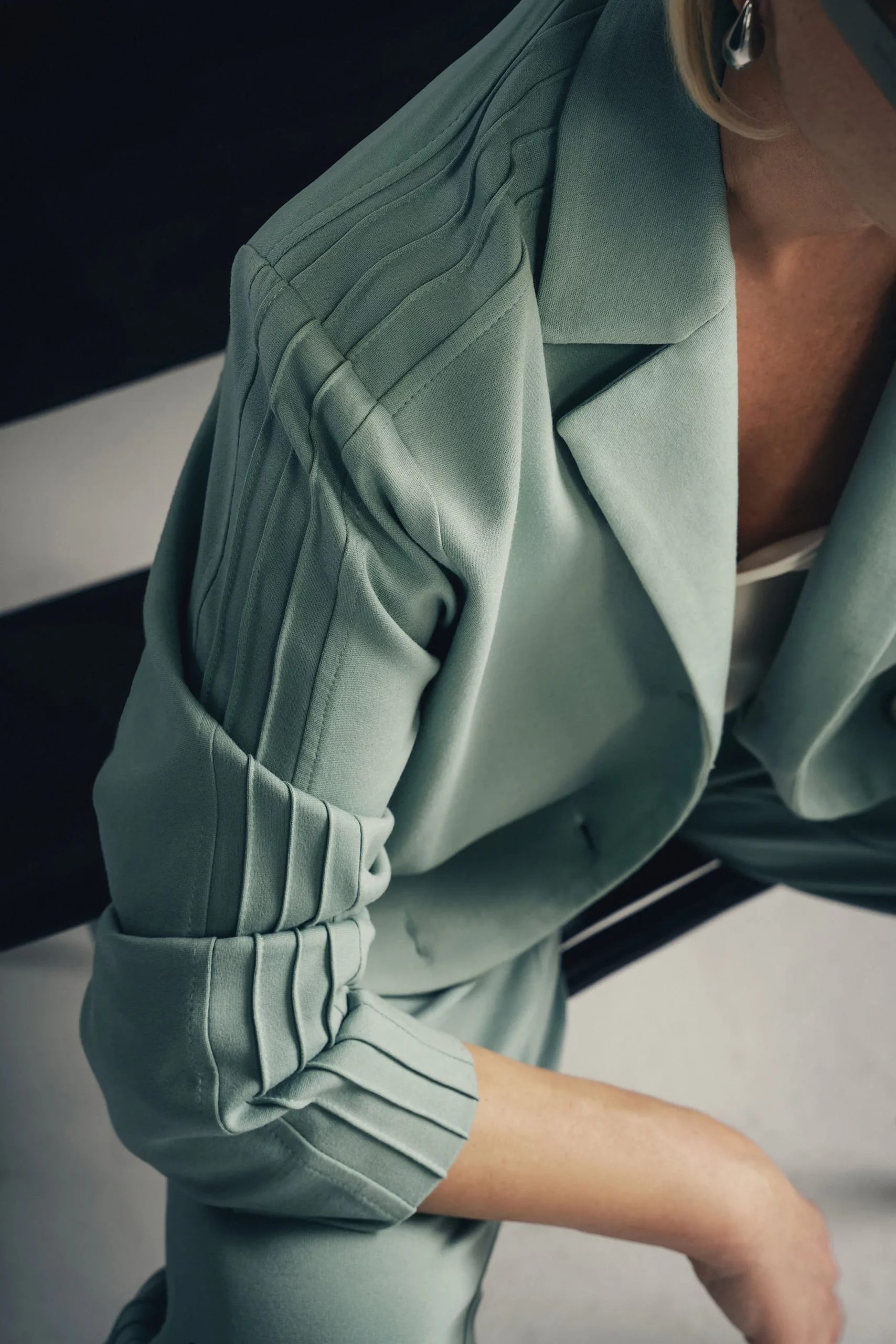 Kobieta siedzi na czarnym krześle, ubrana w zestaw w kolorze szałwi - luźną oversizową marynarkę Jazz z ozdobnymi lampasami na rękawach i dopasowane do marynarki spodnie. Pod marynarką ma biały top na ramiączkach. Stylizacja jest nowoczesna, elegancka i utrzymana w sportowej elegancji