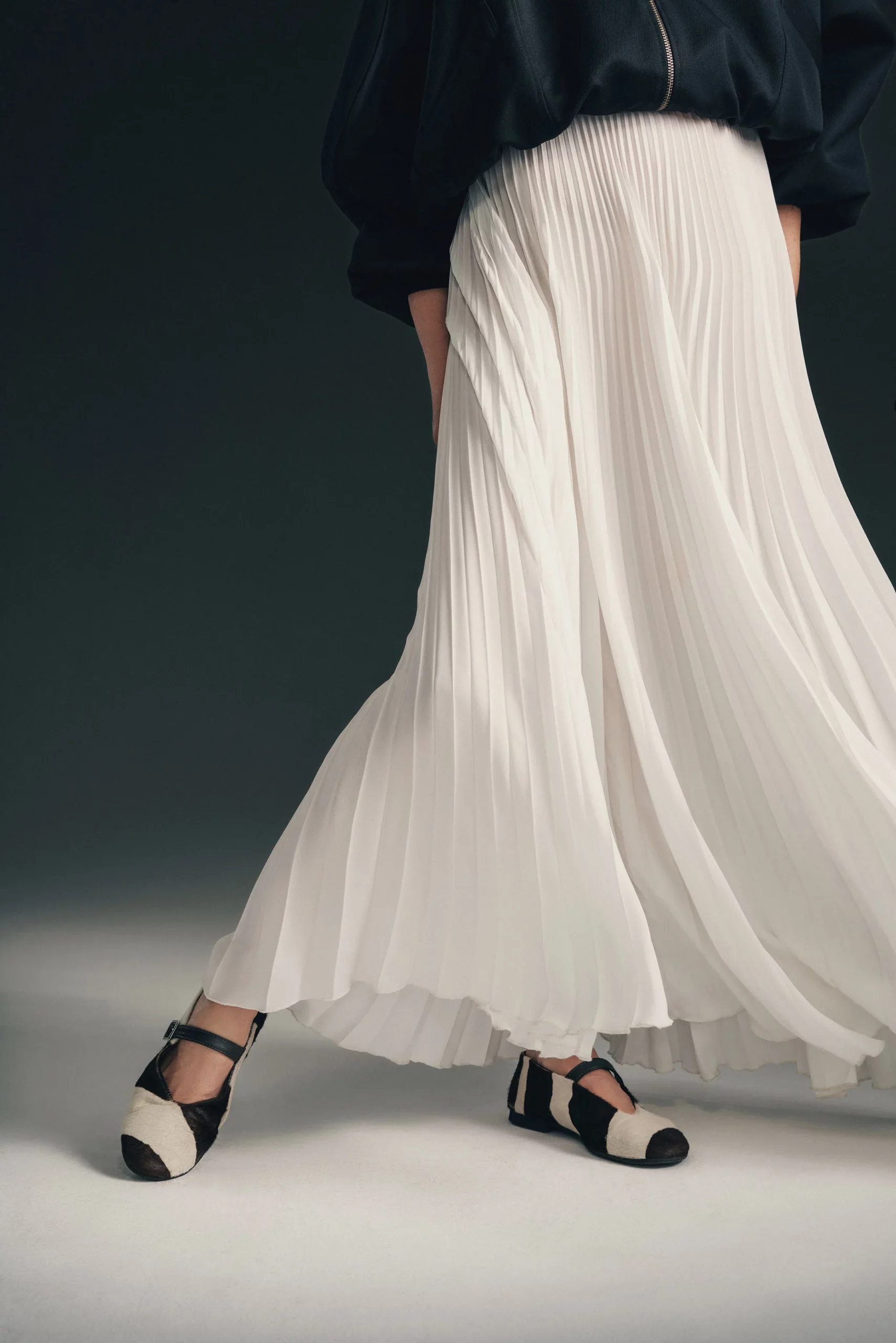 Modelka w długiej plisowanej spódnicy Soleil marki JAK, uszytej z lekkiej, falującej tkaniny, siedzi w minimalistycznej scenerii