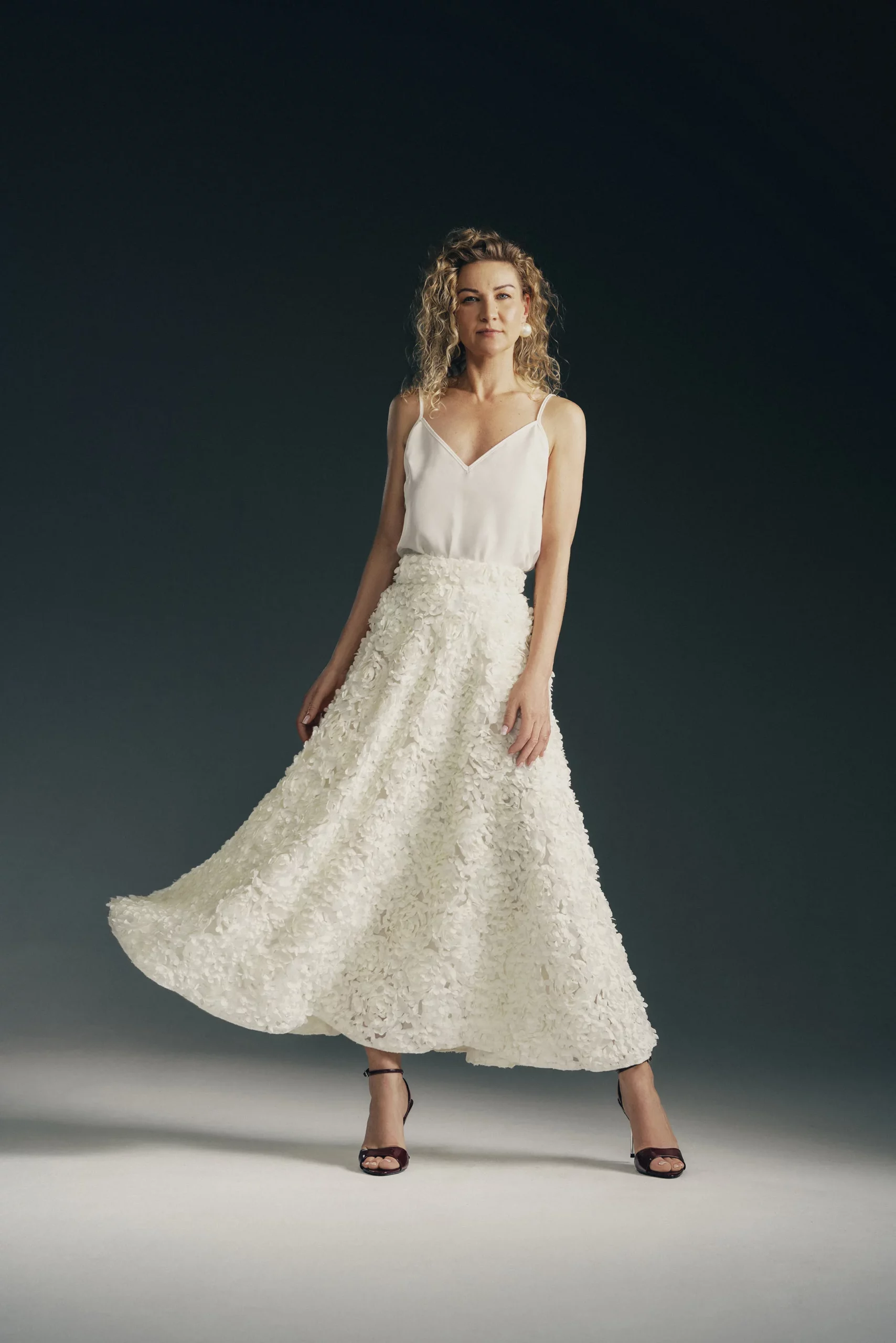 Kobieta ubrana w elegancka spódnice Sun wykonana z lekkiej tkanin 3d w odcieniu ecru. Spódnica o klasycznym kroju z półkoła, z przestrzennymi płatkami i listkami, posiada boczne kieszenie, jedwabną podszewkę i pas podszyty jedwabiem. Stylizacja subtelna, kobieca i wyrafinowana