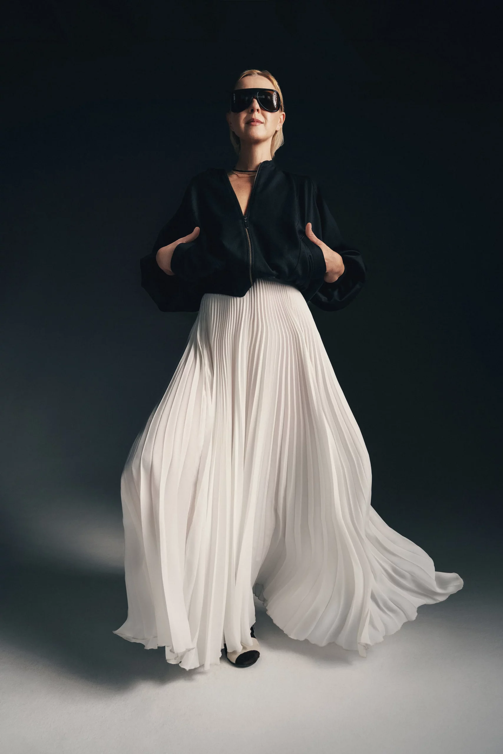 Modelka w długiej plisowanej spódnicy Soleil marki JAK, uszytej z lekkiej, falującej tkaniny, siedzi w minimalistycznej scenerii