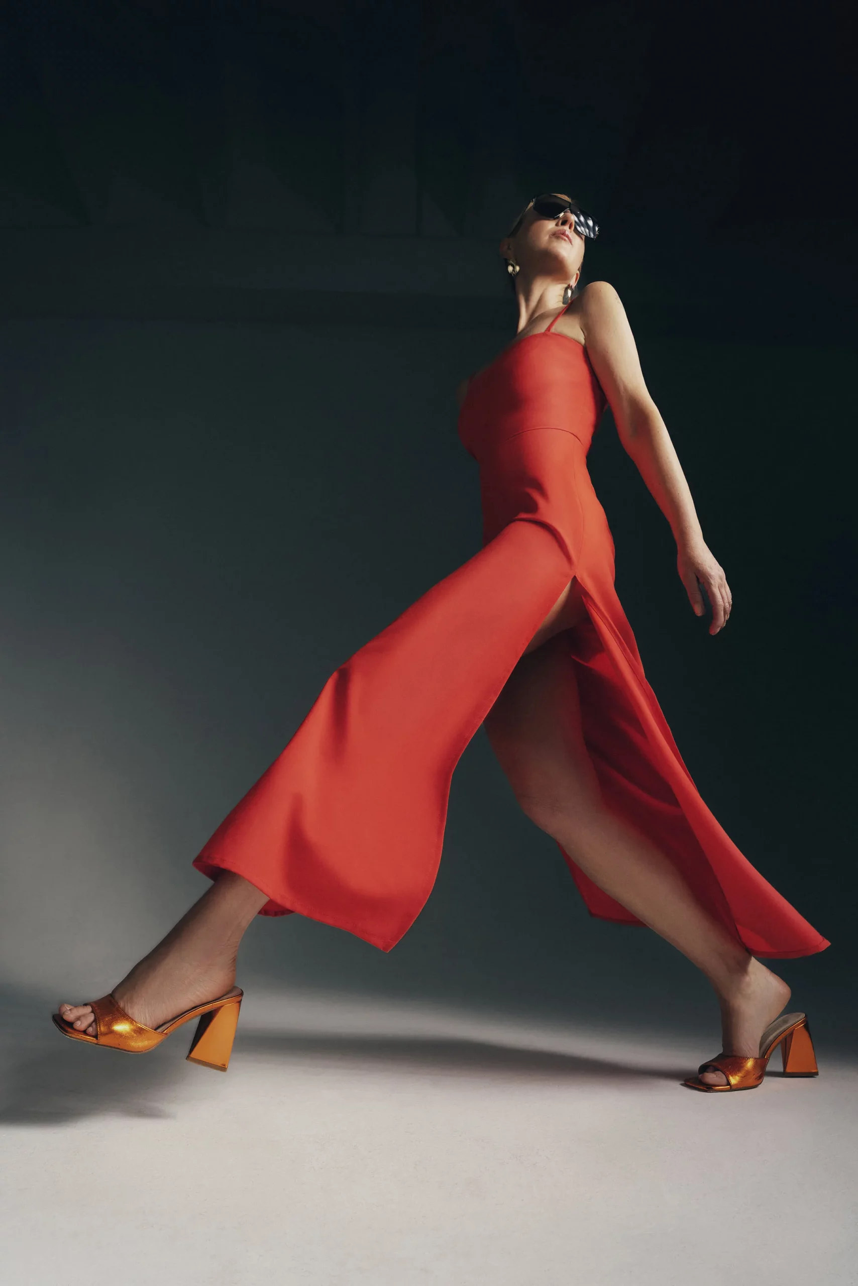 kobieta ubrana w długą sukienkę z lekkiej wełny w kolorze czerwonej pomarańczy, sukienka na cienkich ramiączkach, odcinana w pasie, z głębokimi rozporkami po bokach i podwójną górą, stylizacja lekka, elegancka i zmysłowa